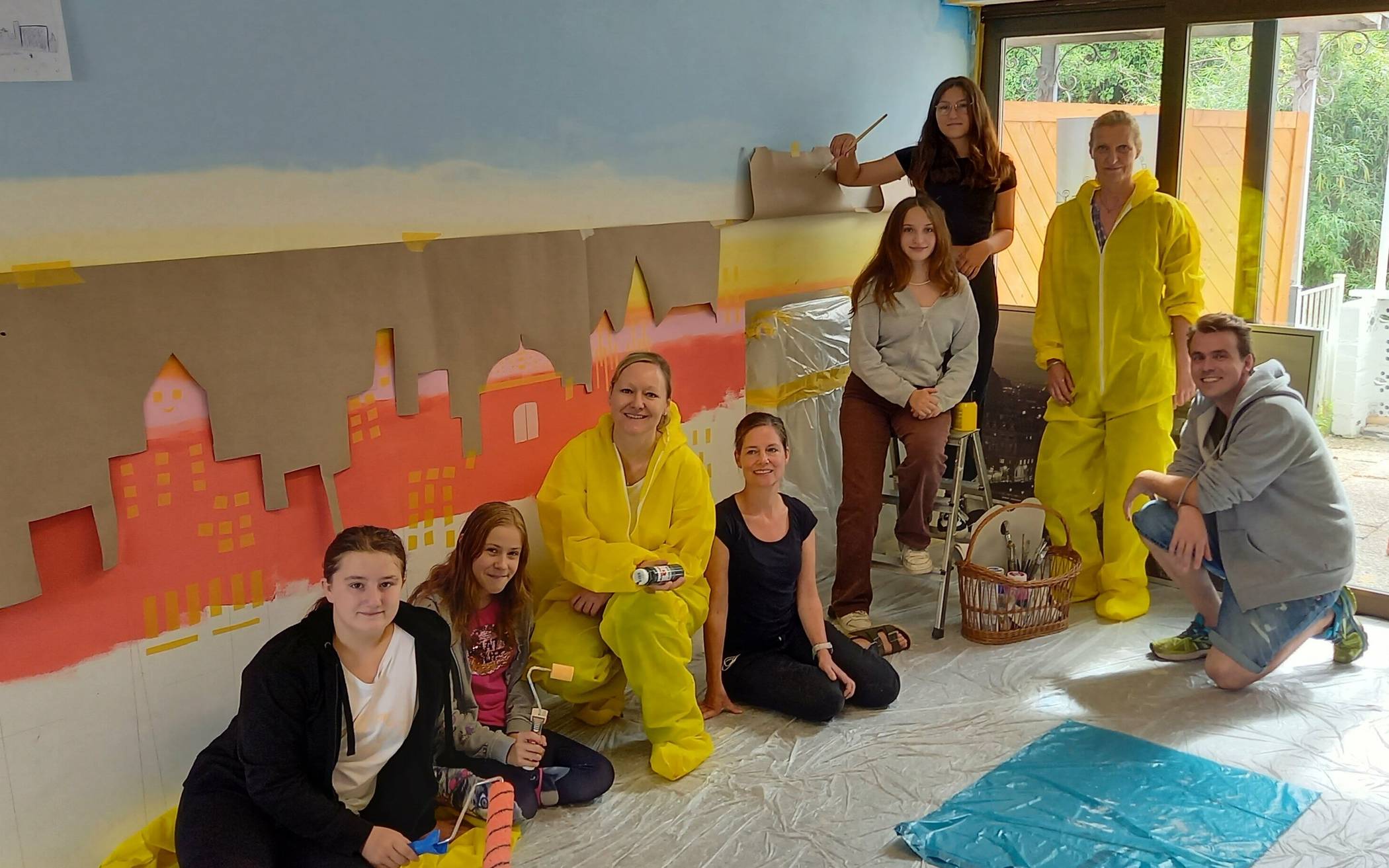 Schulsozialarbeiter Mark Vanck (rechts), Künstlerin Christine Puth (2. von rechts) sowie die Schulsozialarbeiterinnen Nina Focke und Saskia van Hattinga Voß (3. und 4. von links) haben mit Schülerinnen der Robert-Schuman-Europa-Schule im Ferienprojekt das Büro der Schulsozialarbeiter mit einer City-Skyline im Sonnenuntergang verschönert.  