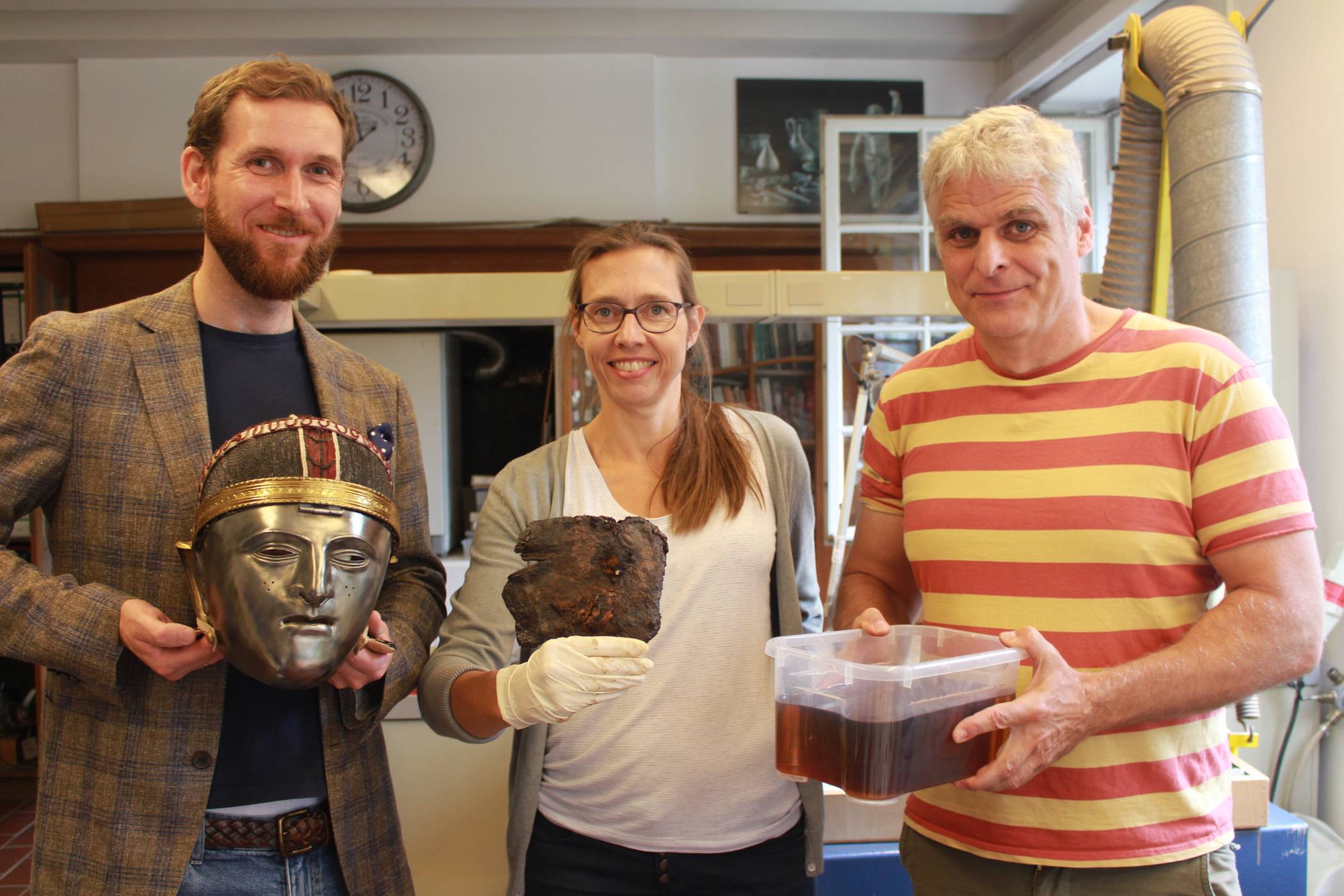  In der Werkstatt des archäologischen Museums Burg Linn: Museumsleiter Dr. Boris Burandt  zeigt die Nachbildung einer römischen Maske, die die Reiterkrieger vom Stamme der Bataver trugen. Das originale Fundstück taucht Restauratorin Eileen Wolff zur Entsalzung in ein Natronbad, das Stadtarchäologe Dr. Hans-Peter Schletter bereit hält. Darin muss es sechs Monate verbleiben. Foto: Müller 