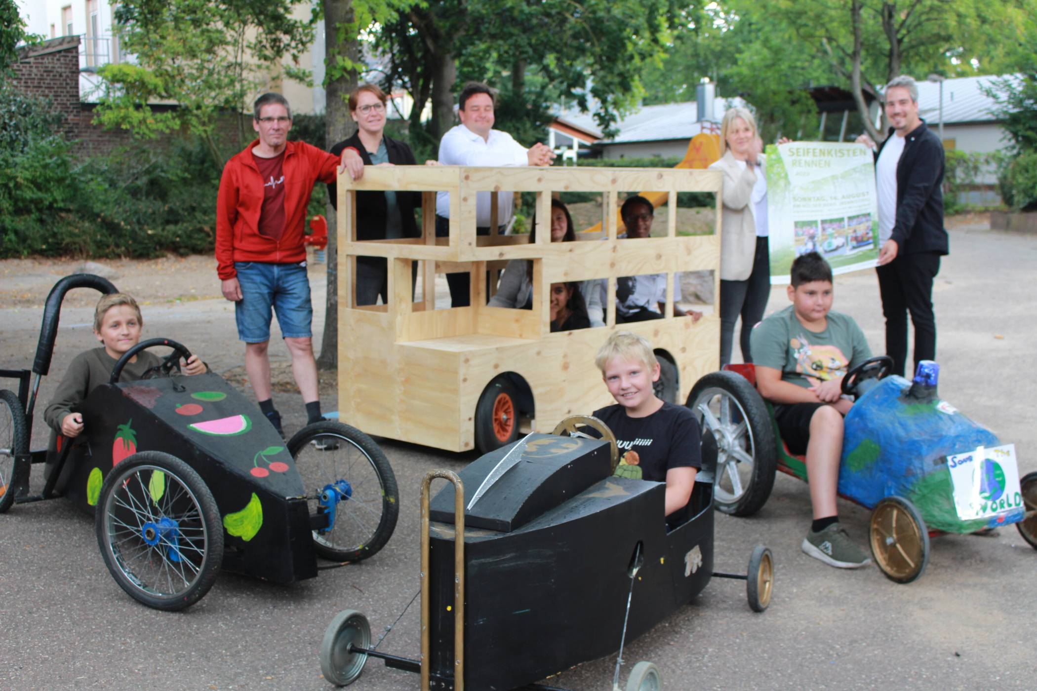 Im Freizeitheim Süd: Die Kinder nehmen die neuen Seifenkisten mit Freude in Beschlag. Heimleiter Norbert Gabriel (l.) hat mit seinem Team ein besonders auffallendes Gefährt gebaut, einen Miniaturbus aus Holz. Diesen bewundern auch Stadtdirektor Markus Schön und die Leiterin der Jugendhilfe Sonja Pommeranz. Organisatorin Anne Caniels und Kollege Matthias Hoeps präsentieren das Ankündigungsplakat vom Seifenkistenrennen. Foto: Müller