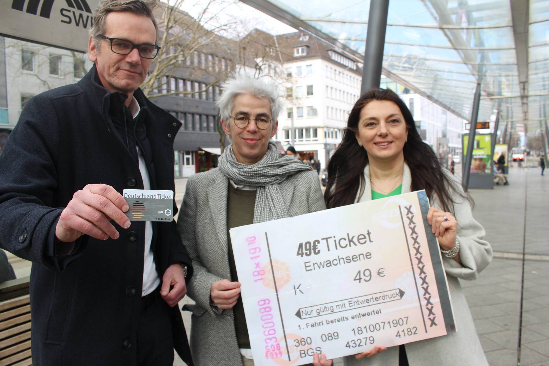 Unterm Glasdach am Ostwall: Bundestagsabgeordnete Ulle Schauws (Mitte) und Landtagsabgeordnete Meral Thoms halten ein Symbolbild des neuen Deutschlandtickets in Händen, SWK-Mobil-Geschäftsführer Guido Stilling hält das echte. Foto: Müller