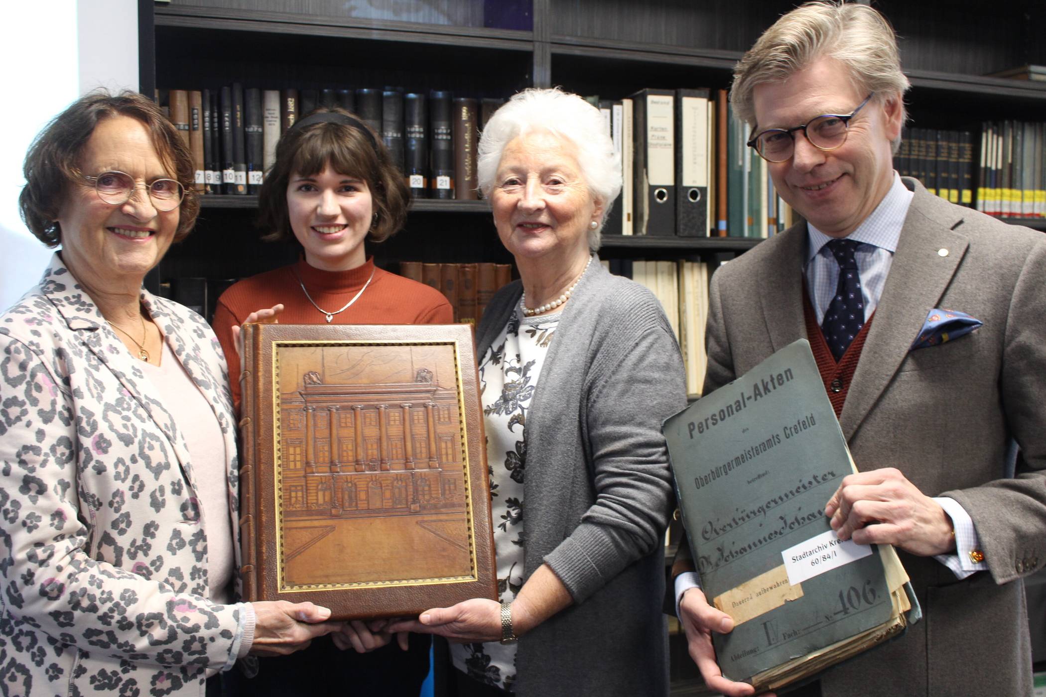 Präsentieren das schwere Fotoalbum mit Coverprägung des Krefelder Rathauses: die Überbringerinnen Renate Rommel-Hohneck (l.) und Roswita Engels (r.) sowie Archivmitarbeiterin  Fenja Schneiders (Mitte). Archivleiter Dr. Olaf Richter hat passend dazu die Personalakte des ehemaligen Buchbesitzers Johannes Johansen aus dem Magazin dazugeholt. Foto: Müller