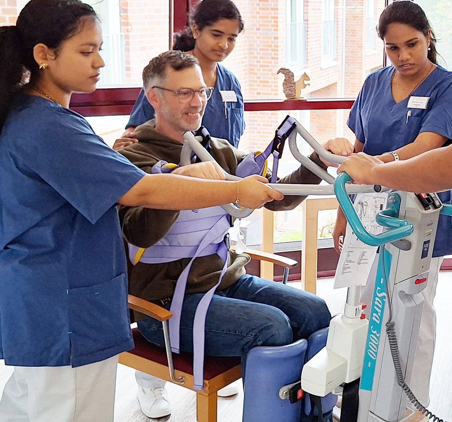  Praxisanleiter Jörg Jeske setzt sich selbst an die Aufstehhilfe, um den indischen Auszubildenden im St. Peter-Stift die Funktion dieses Pflegegeräts zu demonstrieren. 