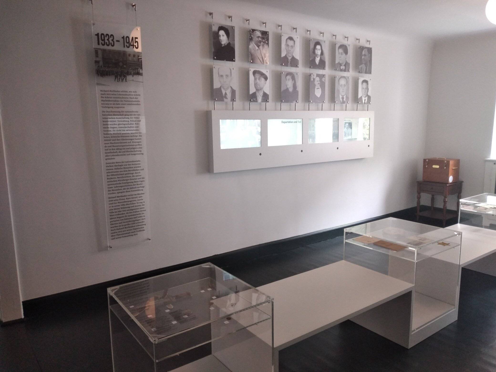 Einblick in die neue Ausstellung: In der Mitte des Zimmers stehen Vitrinen, an der Wand hängen historische Fotos, darunter sind die digitalen Bildschirme mit Erläuterungen angebracht.