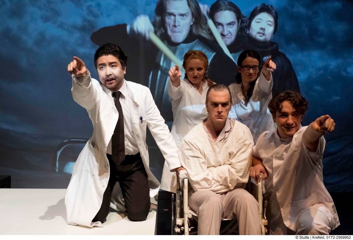 Von Ärzten und Pflegern umgeben: der Komponist Claus Grünberg (Andrew Nolen)landet in der Psychiatrie. Seine Gedankenwelt erscheint auf der Videoleinwand im Hintergrund der Bühne.