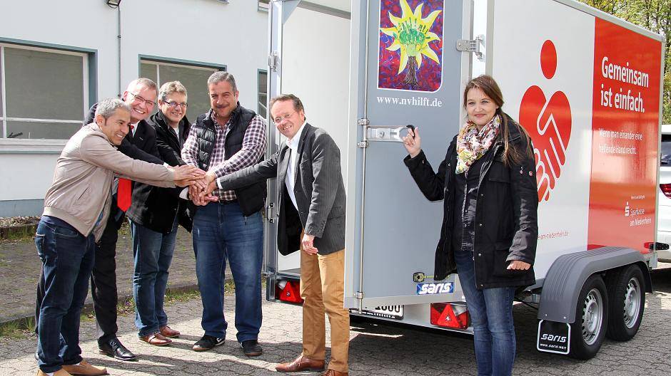 Melanie van der Weiden (r.) präsentiert den neuen Anhänger der Direkten Flüchtlingshilfe, den die Sparkasse mit 2.000 Euro sponserte. Farid Hassani, Bernd Zibell, Torsten Wittrock, Anwar Salem und Klaus Bittmann (v.l.n.r.) freuen sich, dass die Flüchtlingsarbeit in Neukirchen-Vluyn damit effizienter wird.