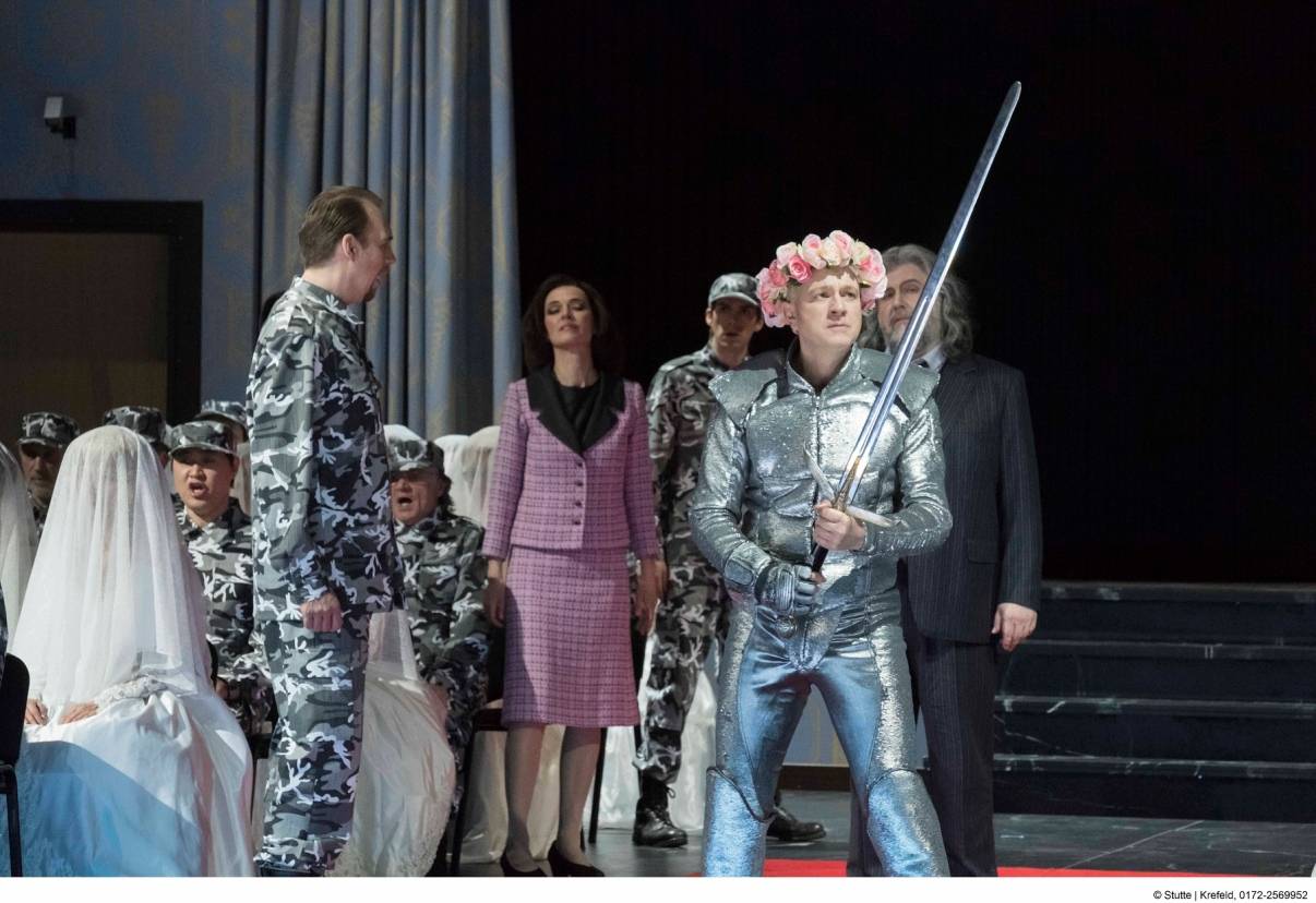 Alle Arten von Kostümen: Wüterich Telramund (Johannes Schwärsky, r.) und seine Frau Ortrud (Eva Maria Günschmann) erscheinen in Designerkleidung, König Heinrich (Matthias Wippich, l.) und seine Krieger tragen NATO-Tarnanzug, die Damen sind züchtig verschleiert und Lohengrin (Peter Wedd) kann sich zwischen silberner Comicfigur und Blumenhippie nicht entscheiden.