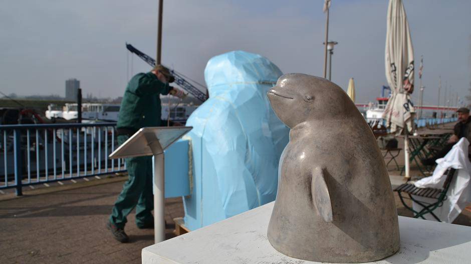 Ein Mini-Rhineheart in Bronze, im Hintergrund Jörg Mazur bei der Arbeit am großen Modell auf dem Ruhrorter Leinpfad. Sechs kleine Bronze-Güsse gibt es bereits, die relativ helle Färbung kommt von einer Wismut-Patinierung, mit der der Kunstgießer experimentiert hat, um der weißen Farbe der echten Belugas möglichst nahe zu kommen. Einen Mini-Rhineheart führt Jörg Mazur immer mit sich, um sich bei der Arbeit daran zu orientieren, ein weiterer — der allererste — wird in der RuhrArt Galerie am Ruhrorter Leinpfad ausgestellt.