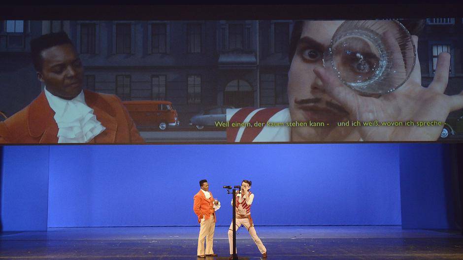 Geteilter Bühnenraum: Unten agieren die Sänger leibhaftig und hautnah, oben erscheinen sie per Videotechnik in laufende Filmbilder eingeblendet. Der Zuschauer mag sich aussuchen, wohin er sein Auge richten will oder ob es hin und herspringen soll.