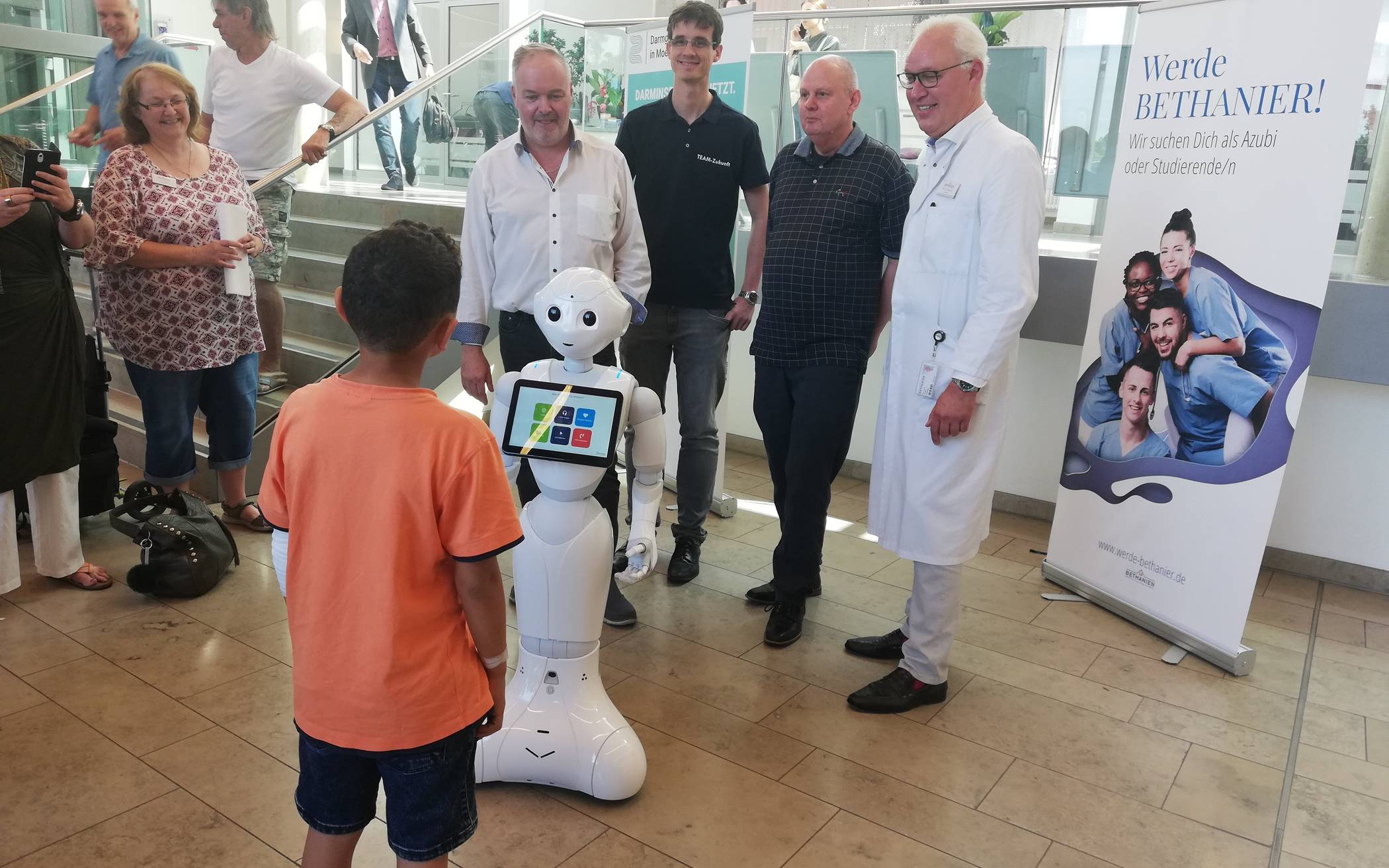 Pepper begrüßt einen kleinen Patienten beim Meet <amp/> Greet im Foyer des Bethanien Krankenhauses: Rainer Becker und Matthias Hofmann (beide ShowBotiXX), Ingolf Rascher (Forschungs- und Praxiszentrum humanoide Robotik und KI in Bochum) sowie Dr. Peter Tönnies, Chefarzt der Frauenklinik und Mitglieder der AG Robotik, stellten den Roboter vor, entkräfteten unrealistische Bedrohungsszenarien und erklärten, wie der Roboter in der Praxis Pflegekräfte unterstützen kann.