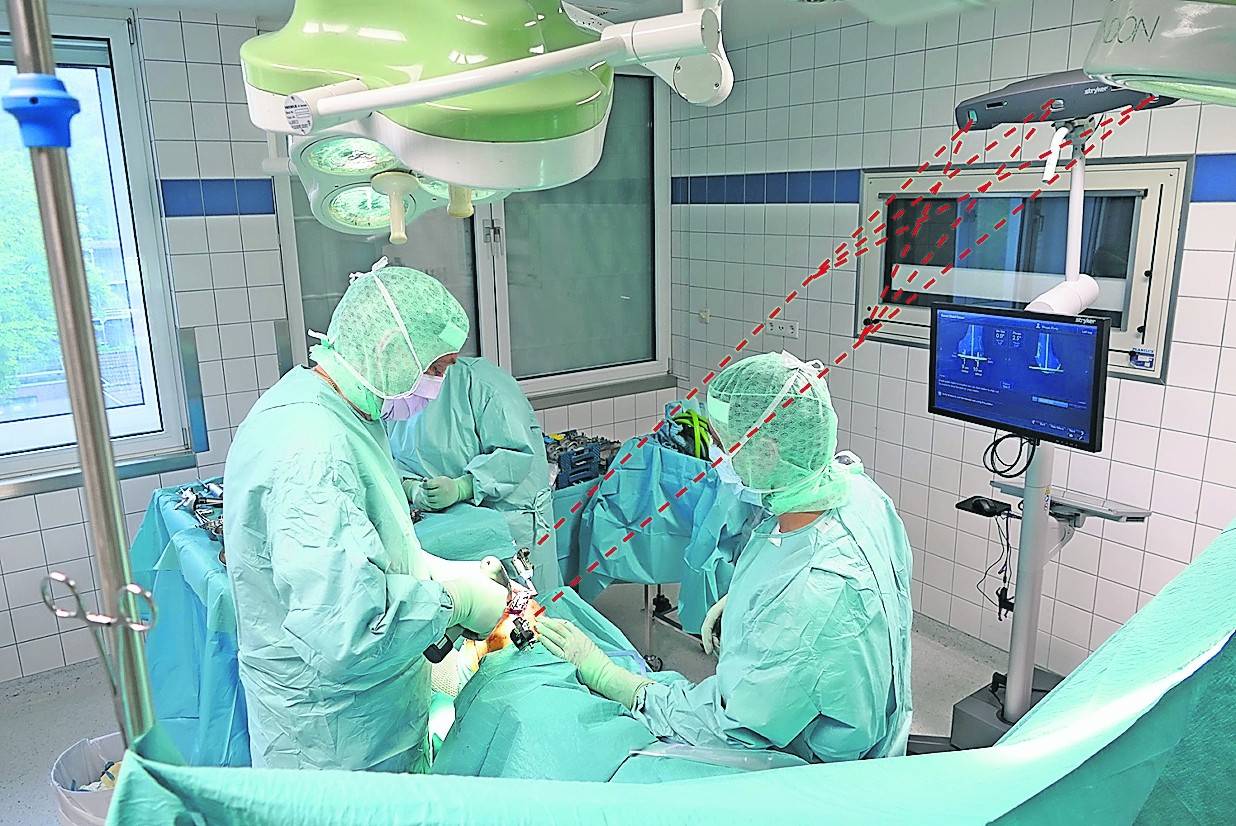 Die Aufnahme zeigt Dr. Martin Fischer (links) bei einer Kniegelenksimplantation (Knie-TEP). Der digitale Operationsnavigator steht mit der Operationsstelle in Verbindung und kontrolliert die Operationsschritte des Operateurs. Für eine anschauliche Darstellung wurden in dem Bild die Infrarotverbindung zwischen dem Navigationsgerät und dem Operationsfeld rot gestrichelt eingezeichnet.