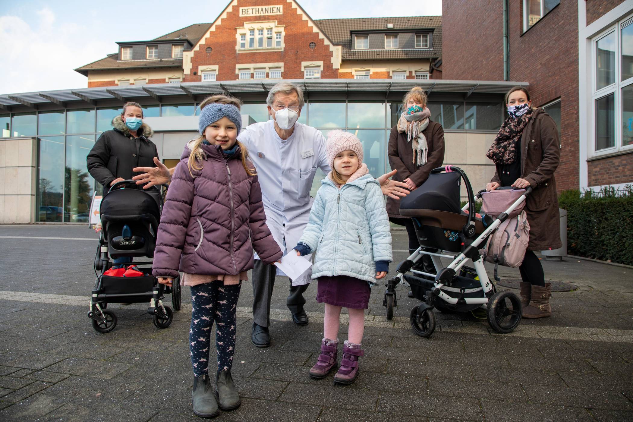 Spendenübergabe vor dem Krankenhaus Bethanien: Mila und Catalina überreichten gemeinsam mit ihren Müttern, Geschwistern und prädagogischen Mitarbeiterin der Kita „Kleine Arche“ sowie der Leiterin des dort umgesetzten Kunstprojektes Stefanie am Weg (2. v. r.) eine Spende an den Chefarzt der Kinderklinik Dr. Michael Wallot.